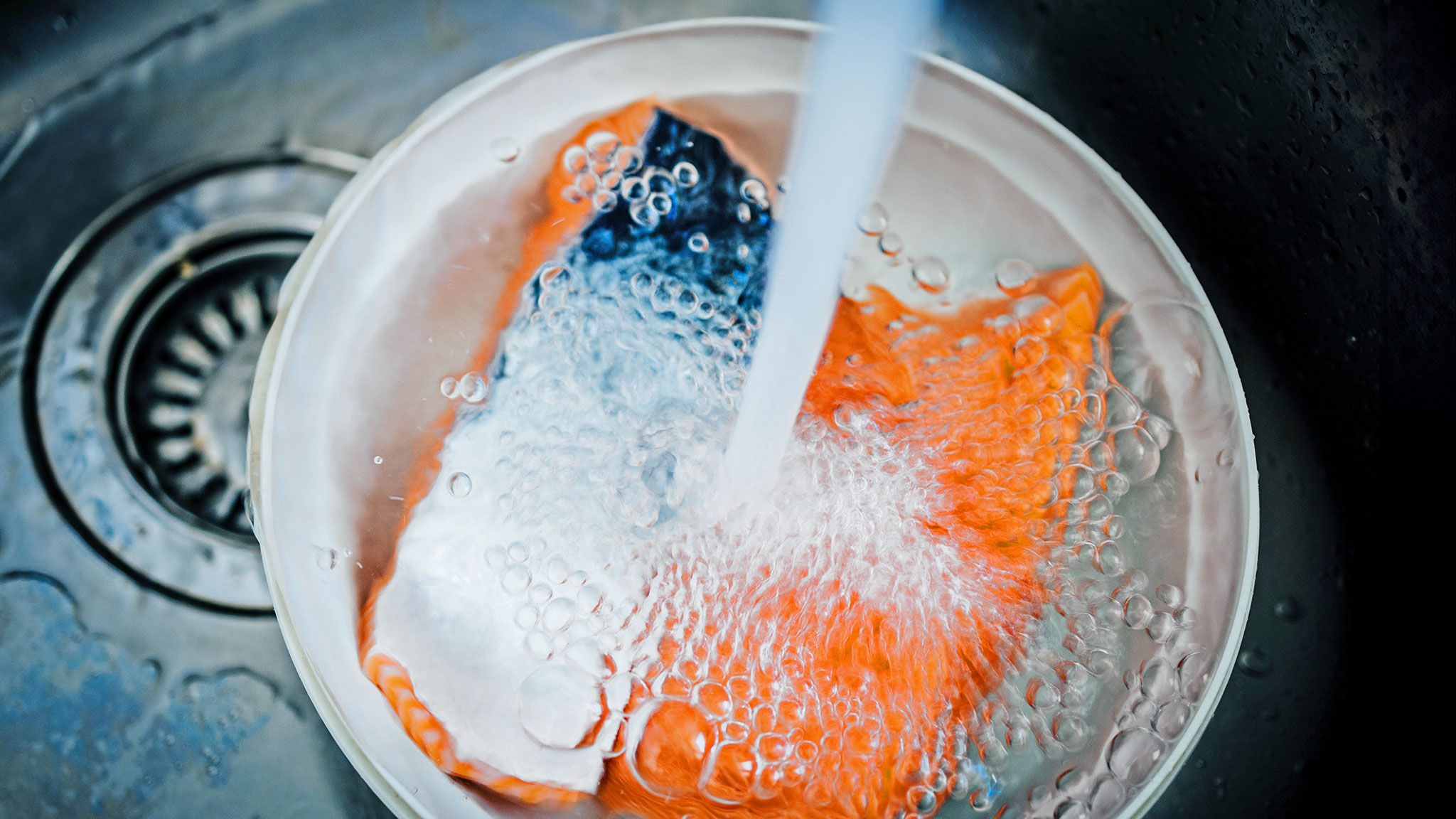 Nahaufnahme eines stählernen Spülbeckens in der Küche, in einer Metallschüssel werden einige Lachsfilets unter einem Wasserstrahl gründlich saubergespült